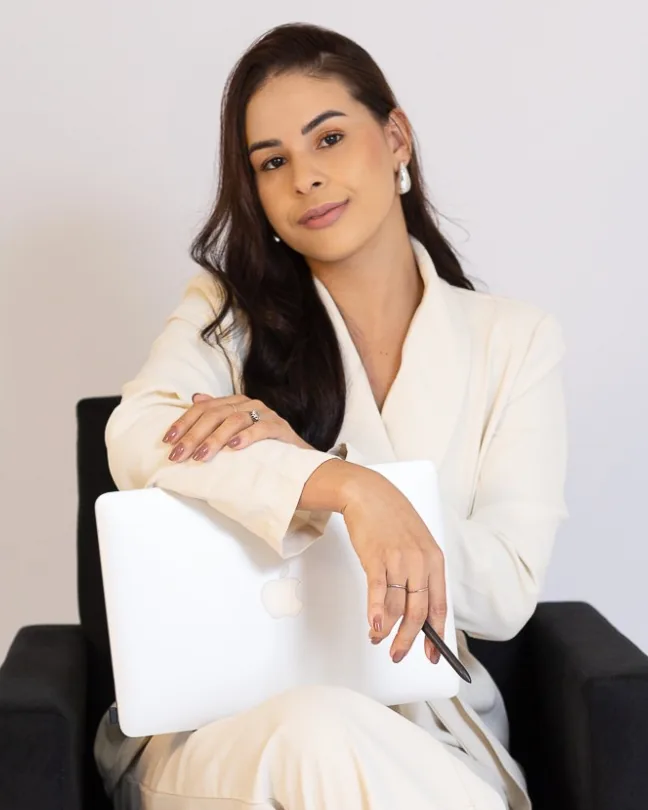 Mislley Santos sentada em uma poltrona preta, de terno creme, com um notebook fechado apoiado no colo e uma caneta na mão direita, olhando para a câmera.