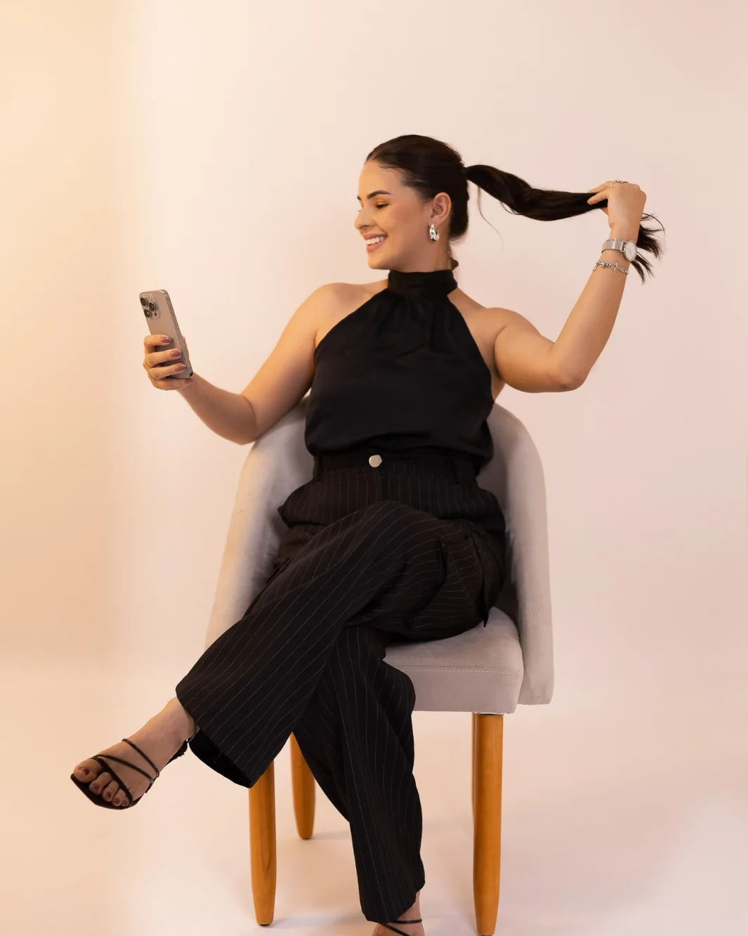 Mislley Santos sentada em uma poltrona, rindo, com o celular em uma das mãos e brincando com o rabo de cavalo.
